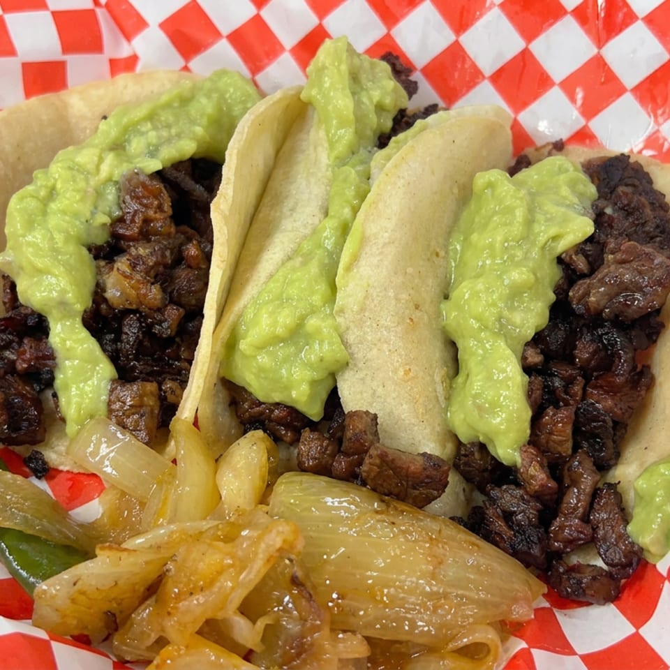 Three corn tortillas filled with grilled meat, topped with guacamole, alongside sautéed onions and green peppers in a checkered basket liner.